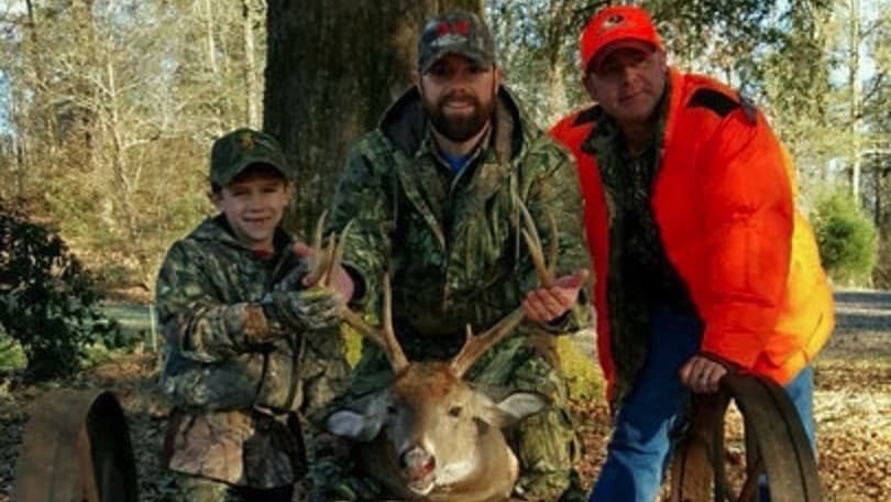 Three hunters with a buck. One kneels holding its antlers, the others flank him. The child smiles, all wear hunting gear.