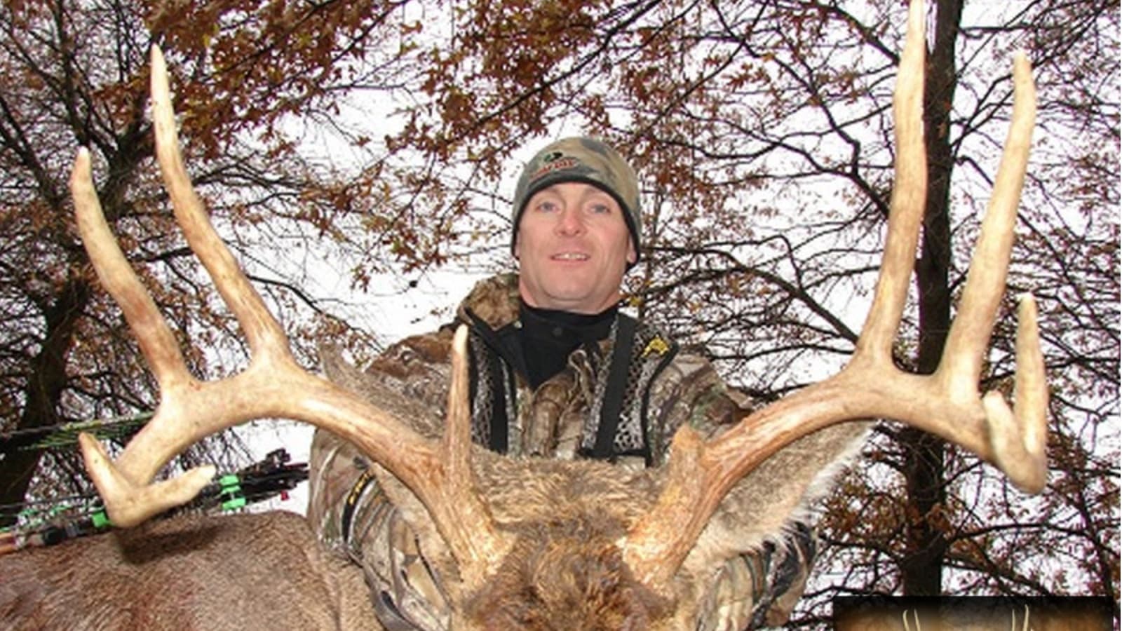 Hunter wearing camouflage holds large buck antlers in front of bare trees.