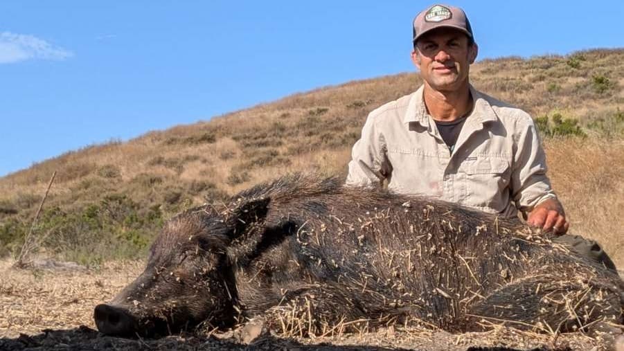 A man smiles next to a large, dead wild boar in a field. Brown hillside and blue sky in background. The boar has thick fur.