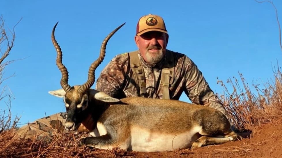 Man in camo with a blackbuck, posing outdoors. The buck lies on the ground, horns curved up. Clear blue sky. Brown earth a...