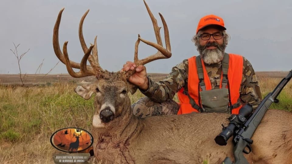 A man in hunting gear kneels beside a large buck, holding its antlers. A rifle is beside the deer. The man wears an orange...