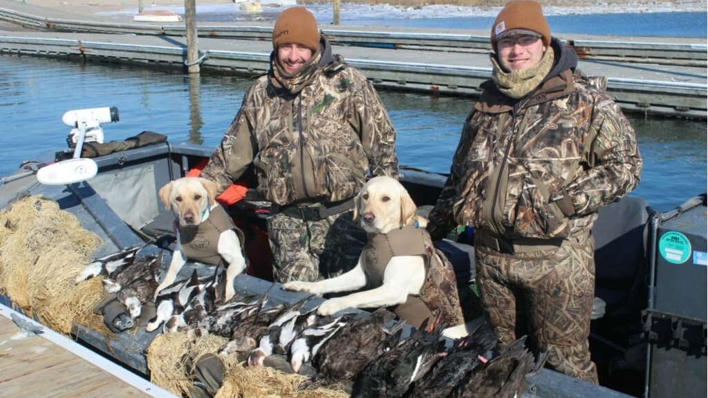 Two men in camo, two yellow labs in vests, and ducks in a boat on water. Smiling men with hunting gear pose with dogs & hunt.