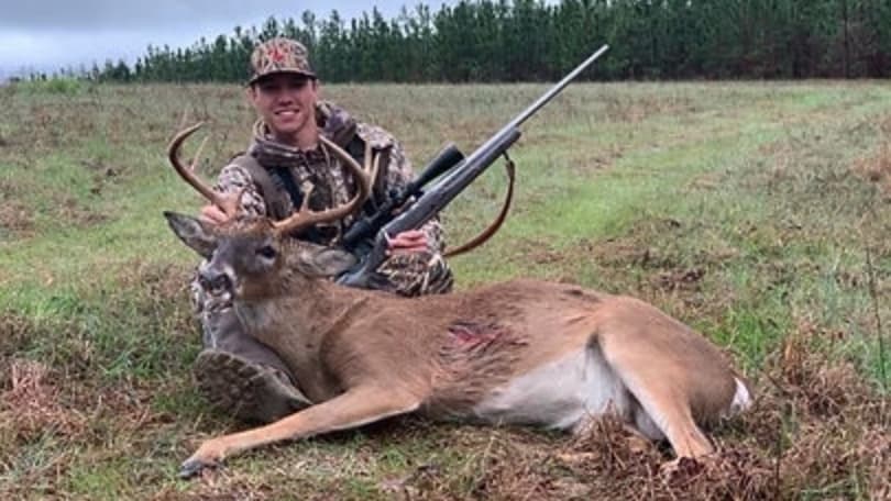 Man in camo holds rifle and poses with a dead deer in field. Deer has antlers and a visible wound. Overcast sky and trees ...