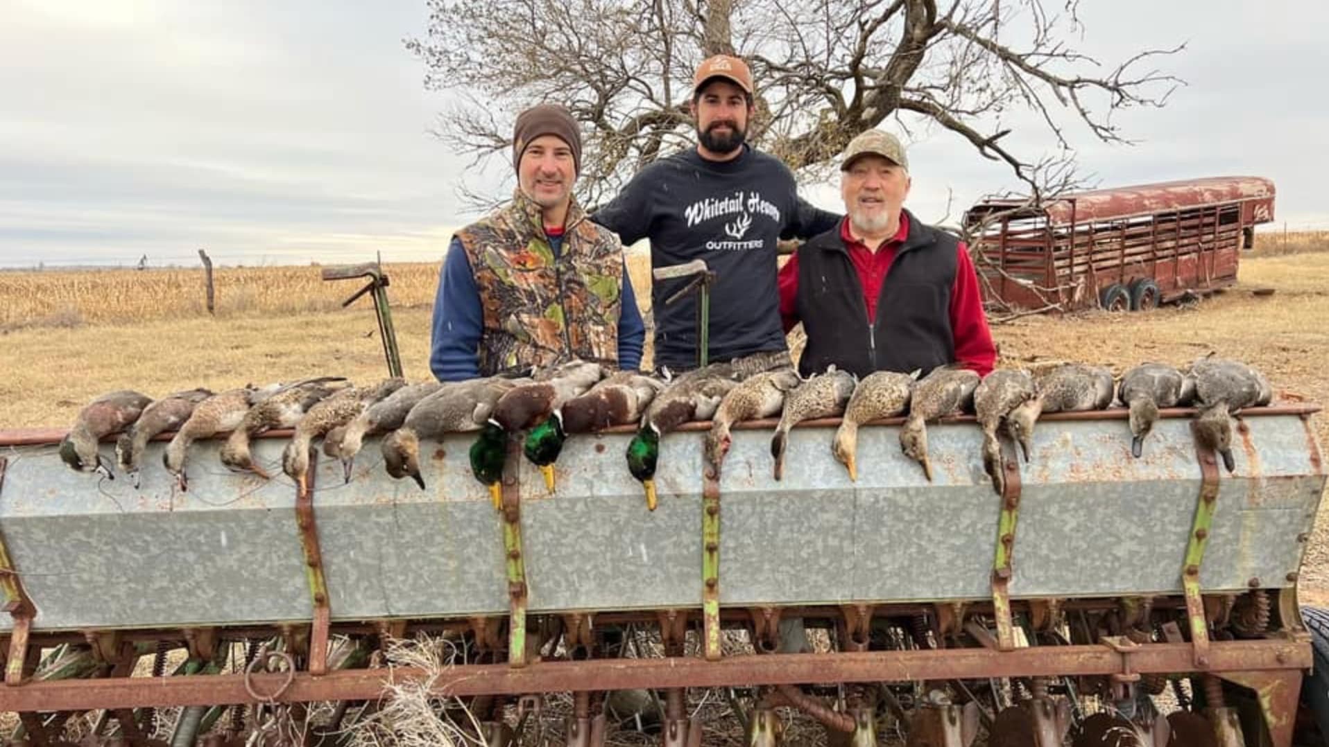 Three men pose with harvested ducks laid out on a planter. Ducks are dark with some green heads. A rusty trailer and tree ...
