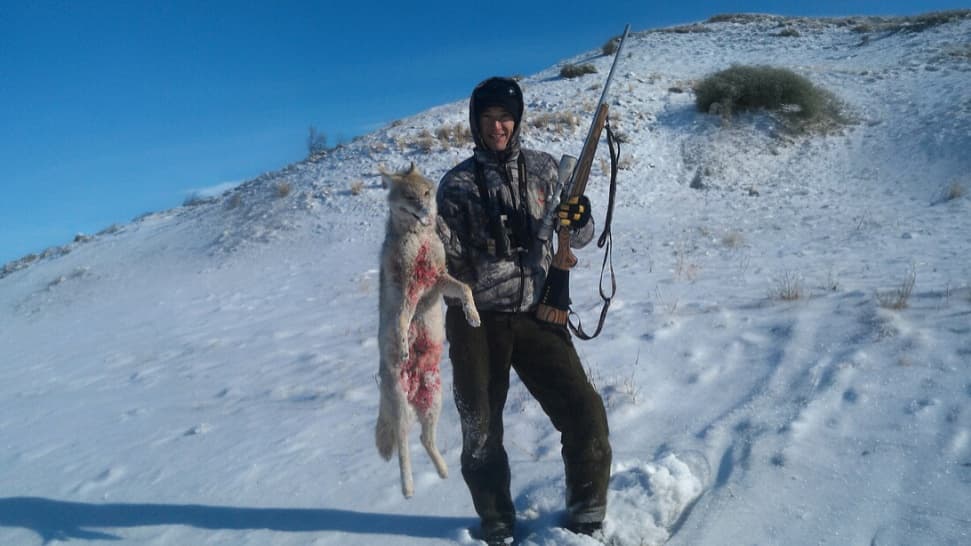 Hunter in camo holds a rifle and a dead coyote, bloodied, in snowy landscape. Blue sky.