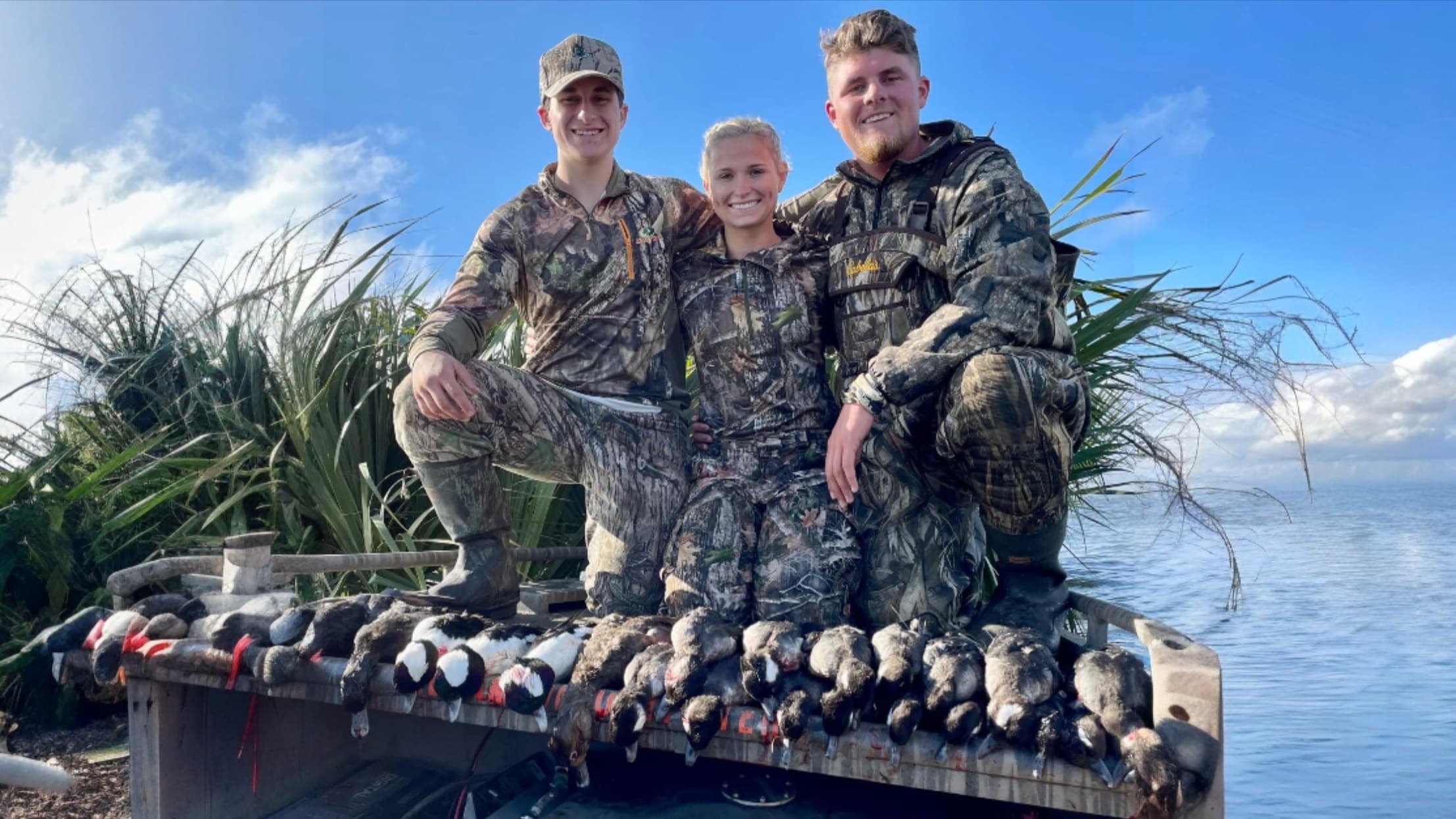 Three people in camouflage pose with a pile of ducks on a boat. The blue sky and water are visible.