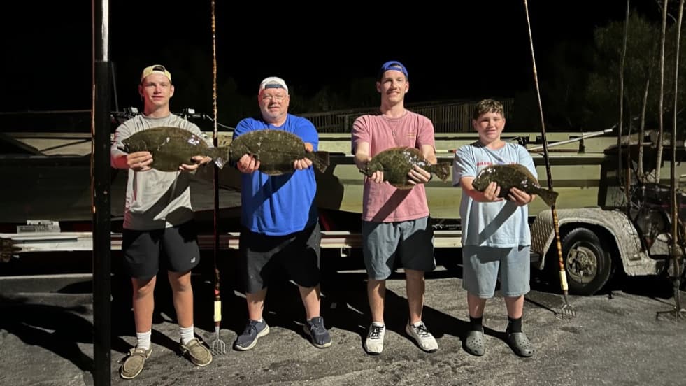 Four people holding flounder fish they caught. Each person stands in a line with a fish, some sporting caps and shorts. Ba...