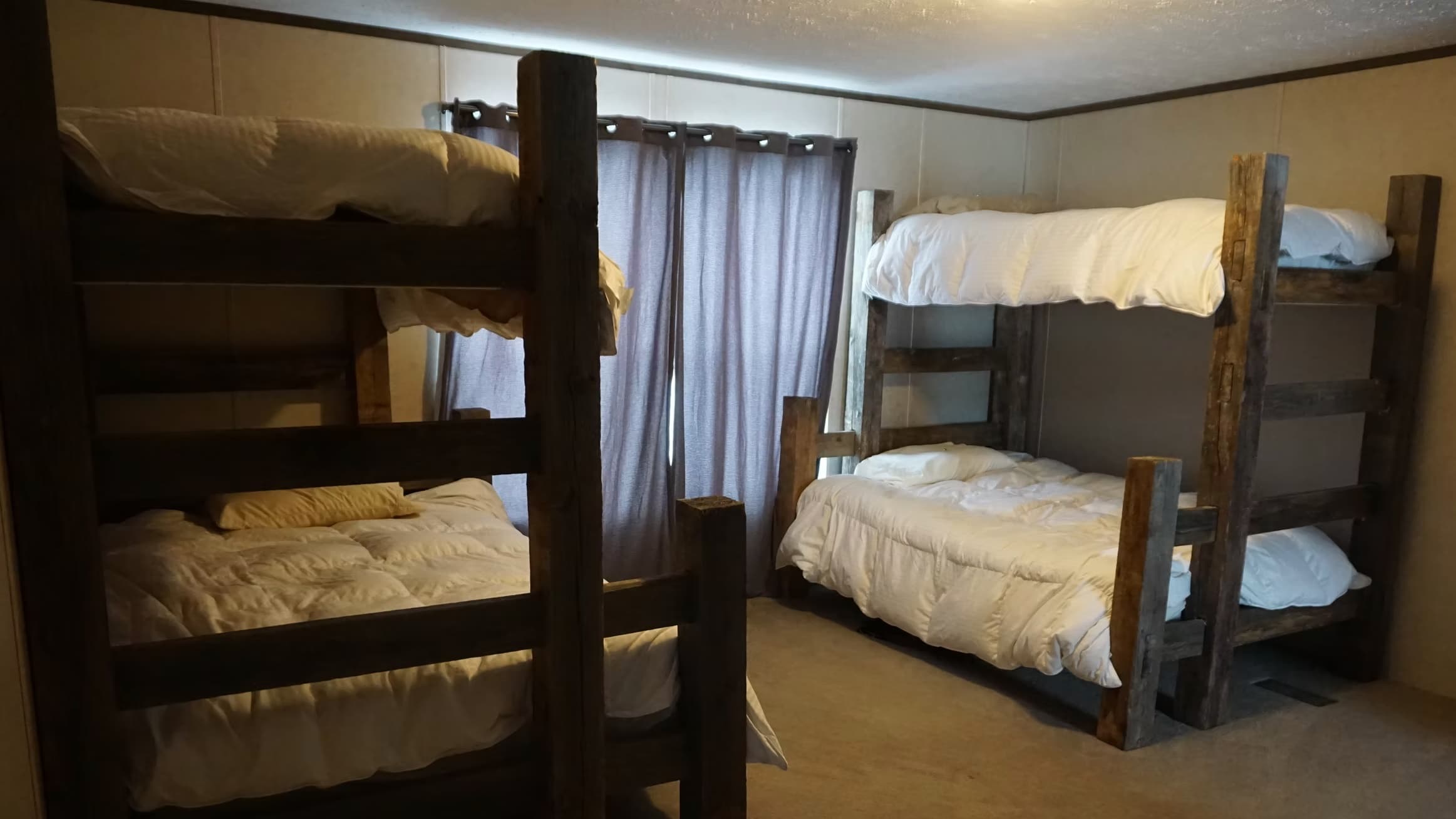 Two sets of rustic wooden bunk beds in a room. Beds have white bedding. Curtains hang between the sets. Dim, warm lighting.