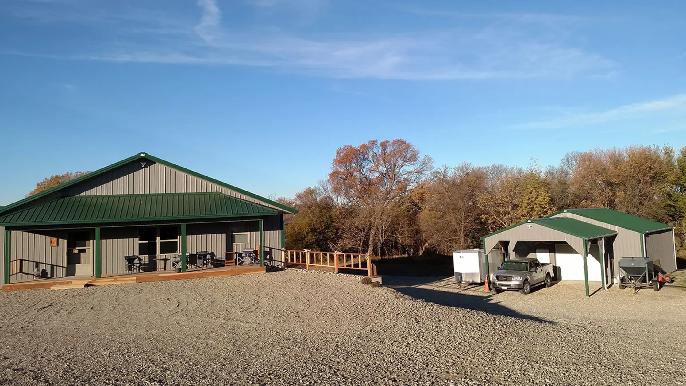 Two metal buildings with green roofs on a gravel lot under a blue sky. One building has a porch. A truck parked in a carport.