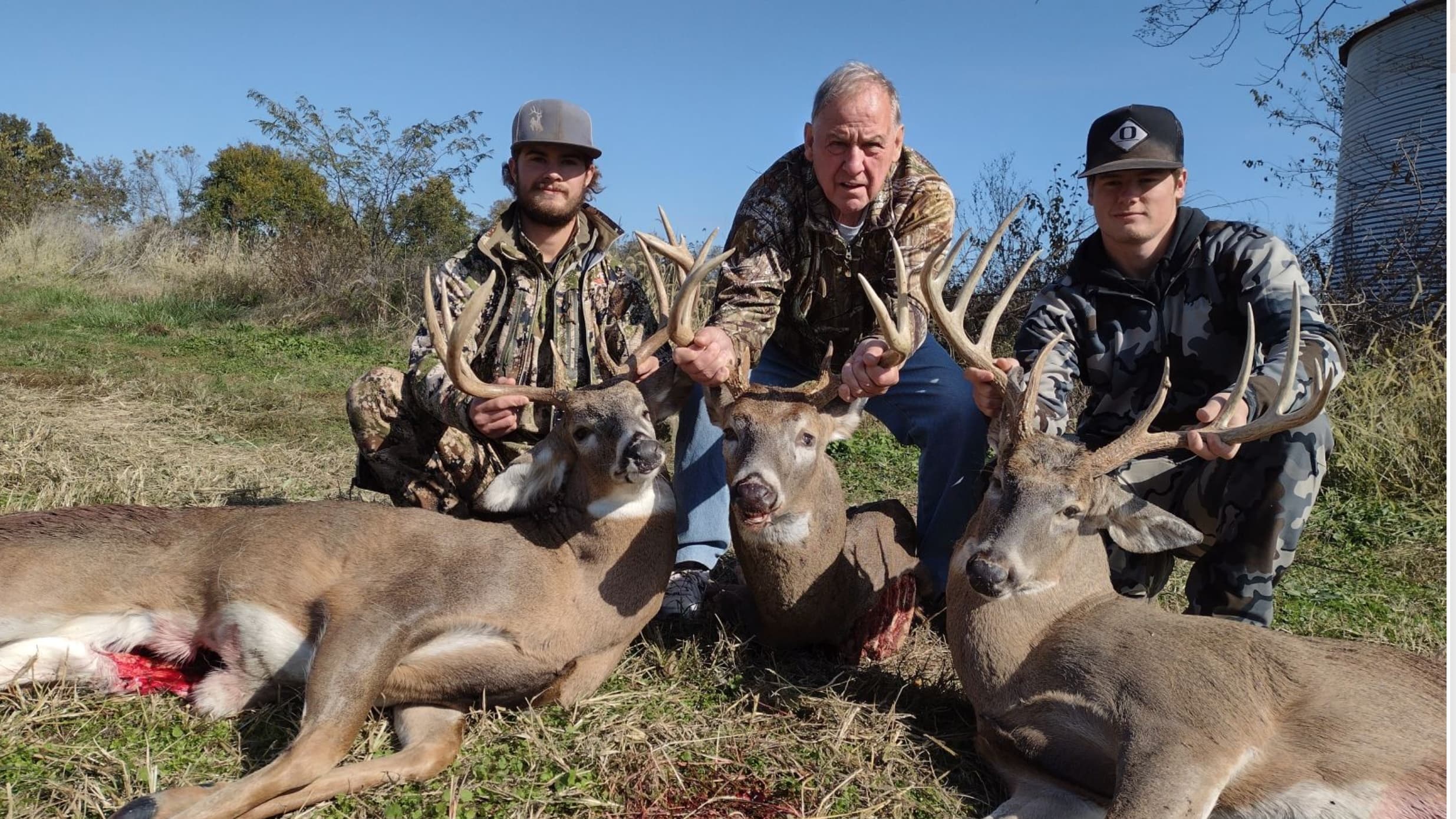 Three hunters pose with their harvested deer outdoors on a sunny day.