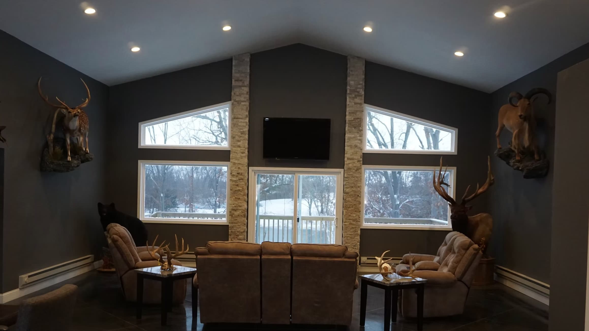 Living room with taxidermy: elk, ram, and deer heads. Dark gray walls, windows overlooking winter landscape. Leather chair...