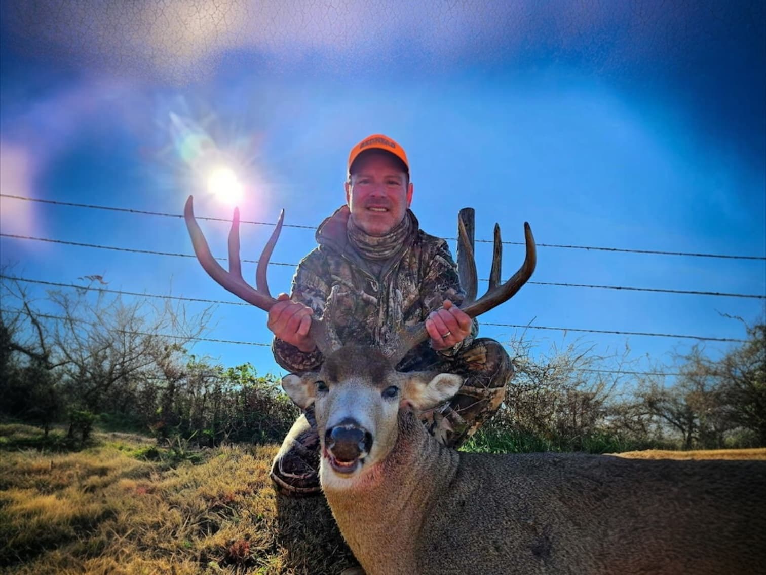 Hunter in camo gear kneels behind a large buck, holding its antlers. Bright blue sky with sun flare. Barbed wire fence in...