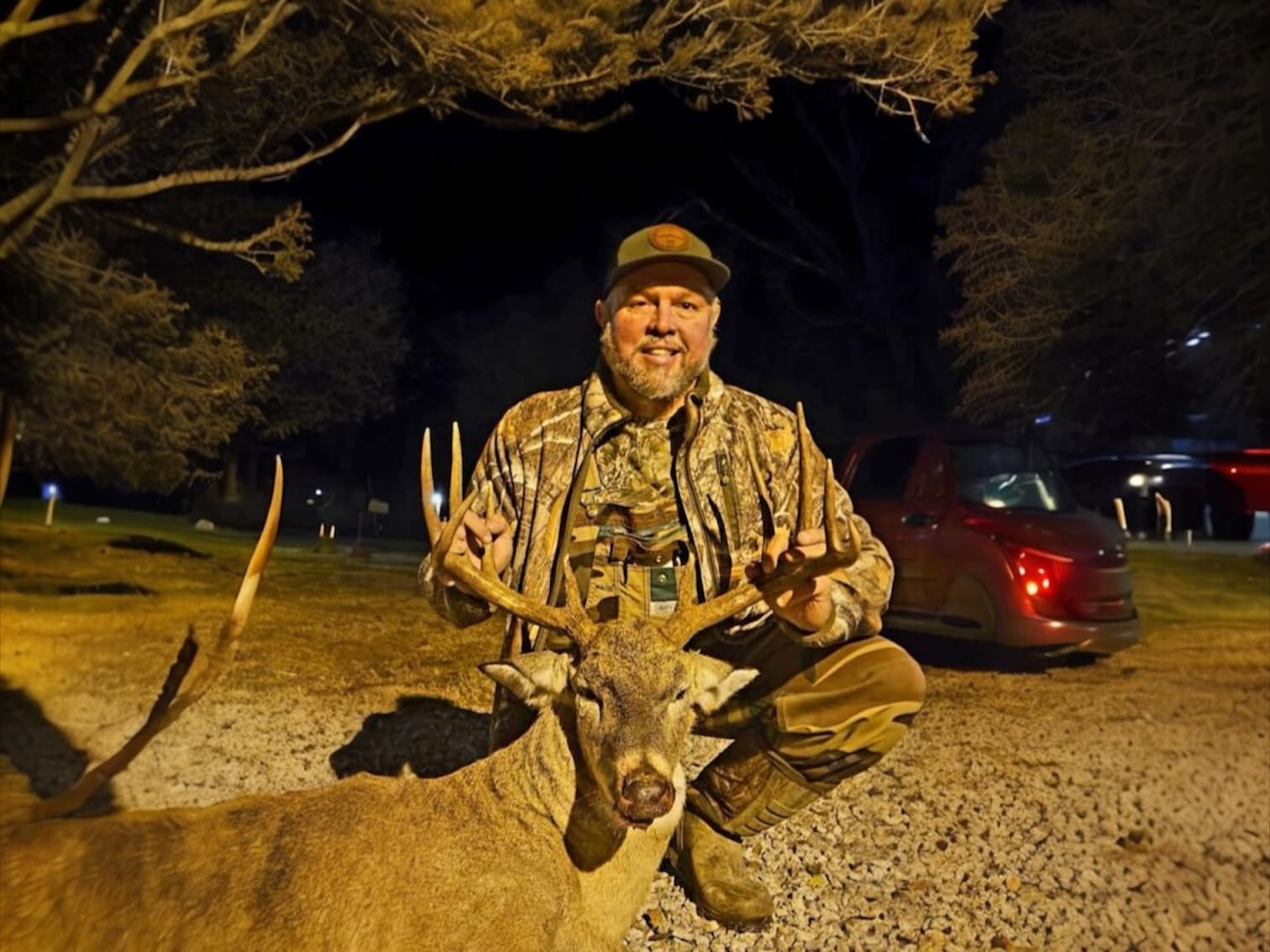 Man in camo kneeling beside a deer, holding its antlers. Night scene with red truck in background.