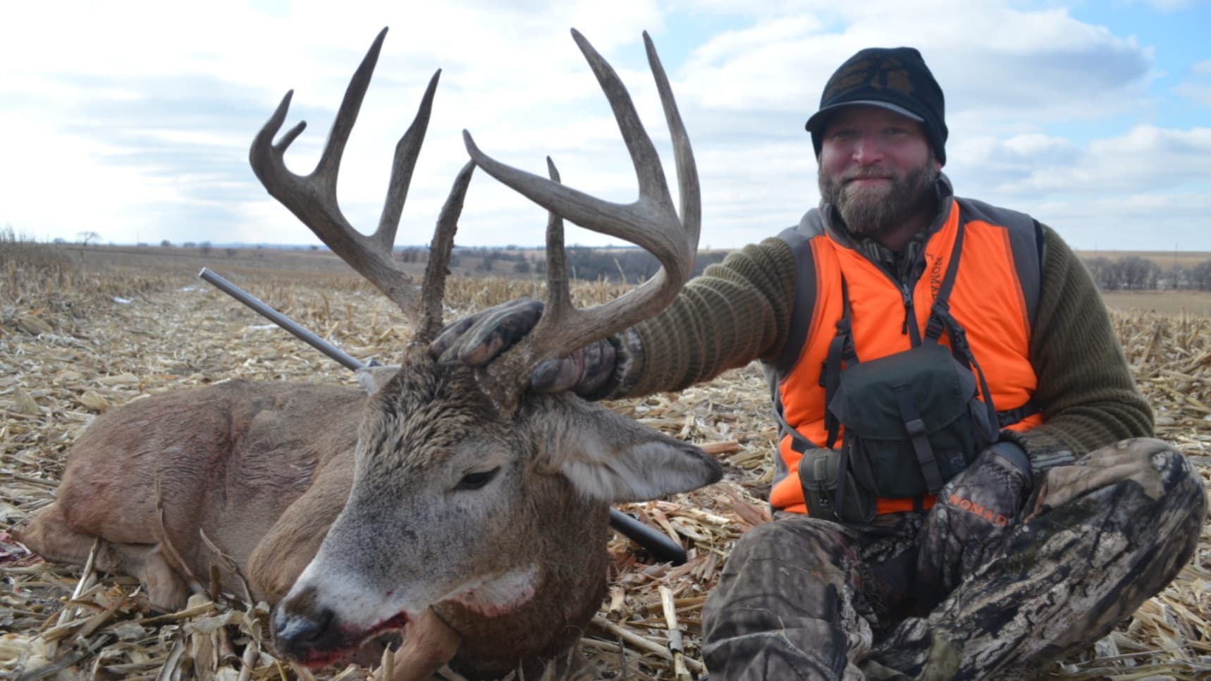 Hunter kneeling beside a large buck with antlers. The hunter wears camo pants, orange vest, and a black hat. Rifle visible...