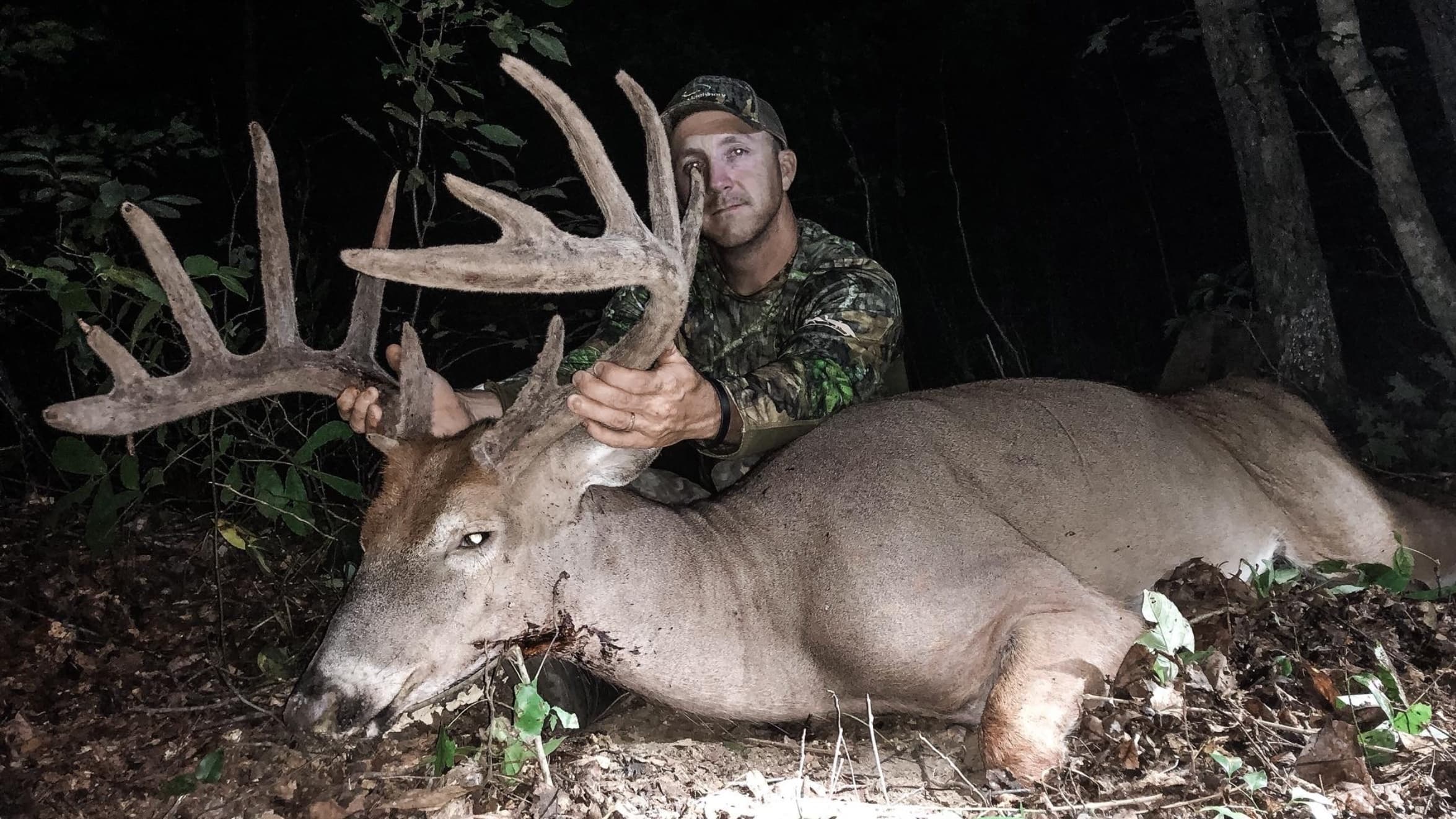 A man in camo holds the large antlers of a fallen deer. The deer lies in fallen leaves and foliage. The man is in a forest...