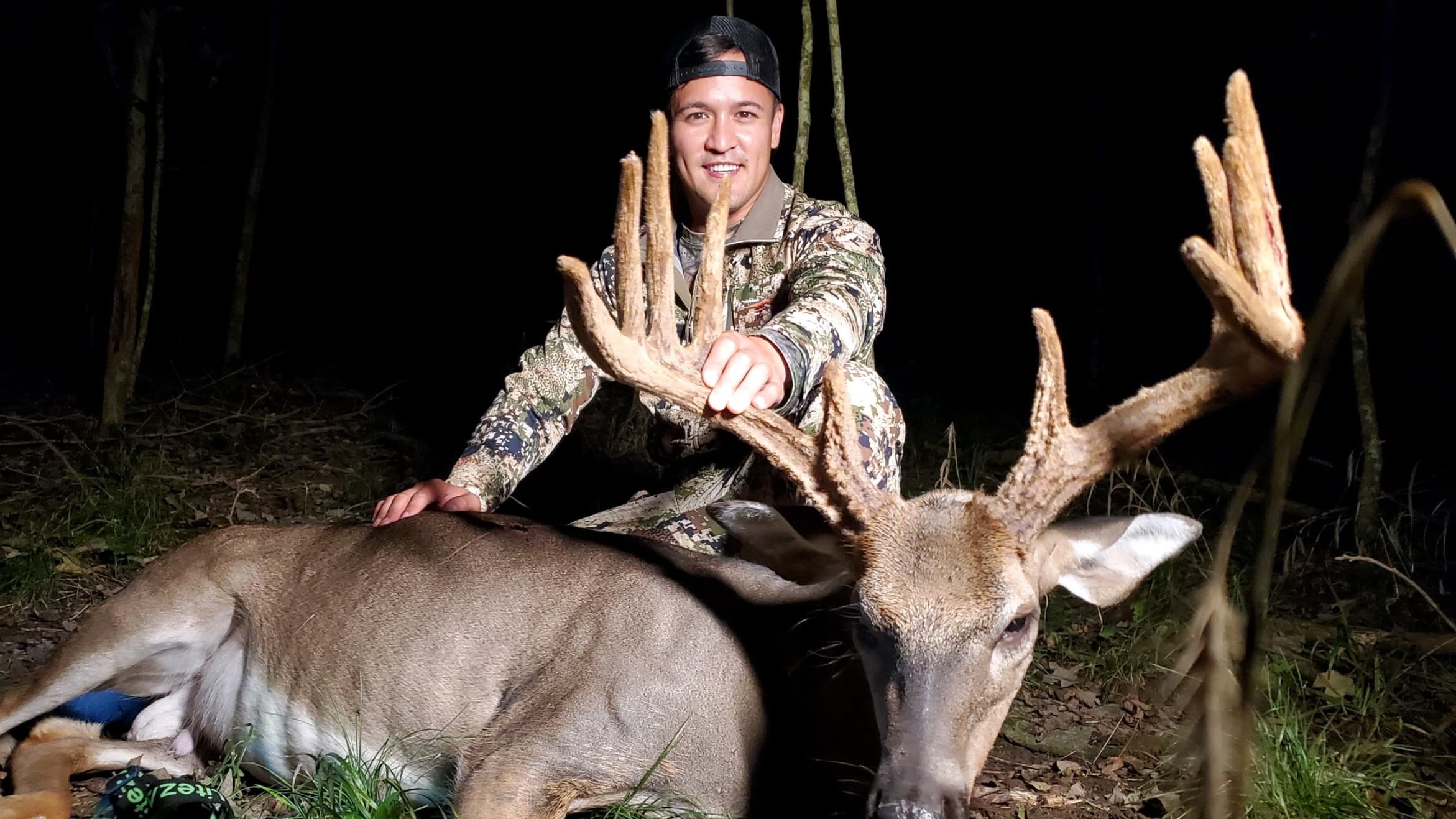 Man in camo holds deer antler, smiling at the camera. A dead buck lies on the ground with the man. Forest at night in bac...