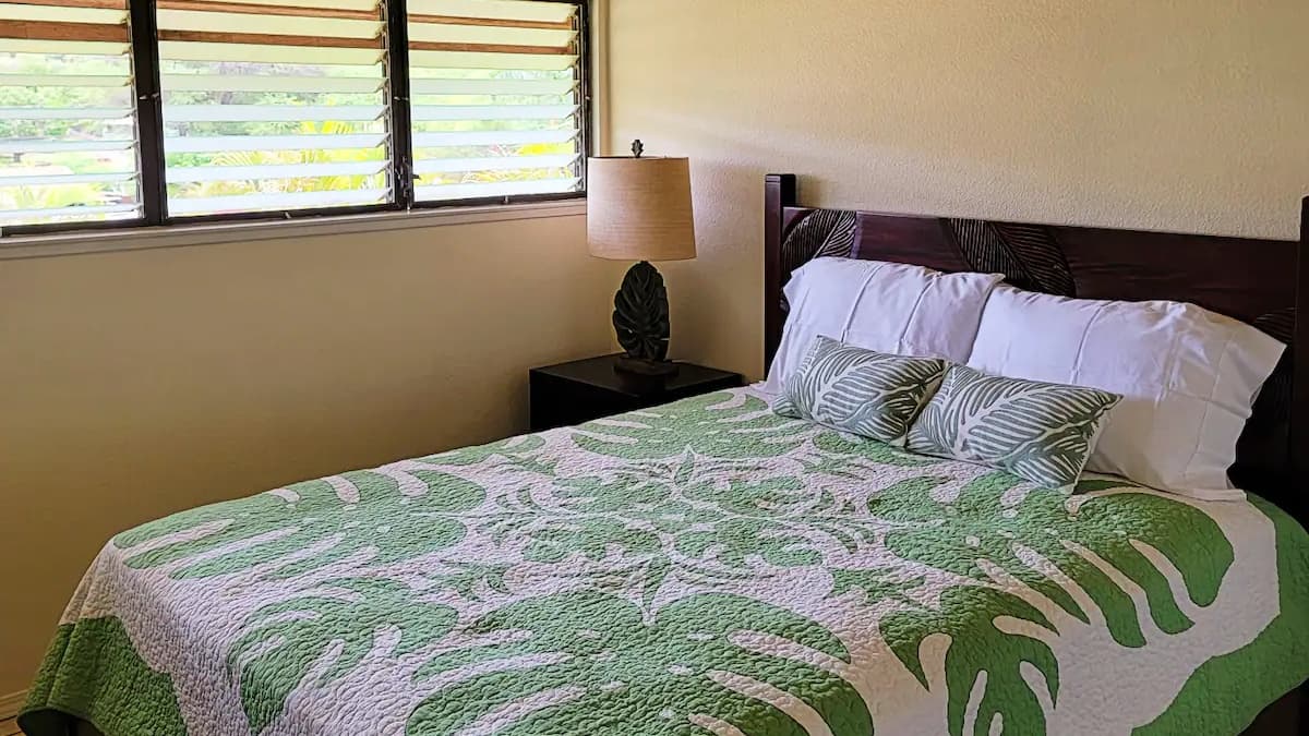Bedroom with a bed. Green and white leaf-patterned quilt and pillows. Dark wooden headboard and nightstand with a leaf-sha...