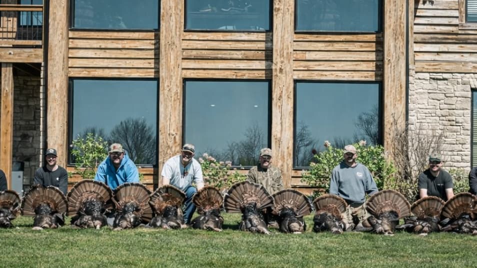 A group of men kneel behind several harvested turkeys, displayed on a lawn in front of a large house. The men pose for a p...