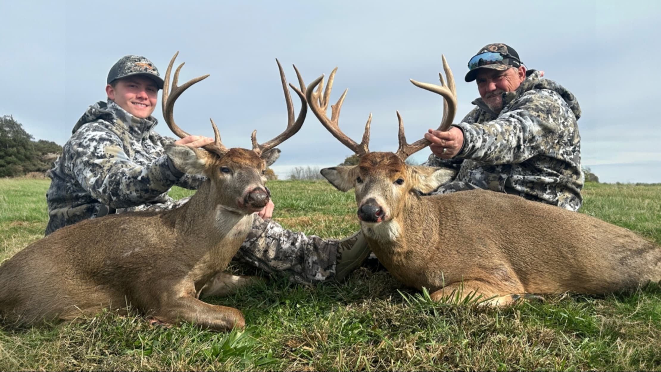 Two men with camouflage clothing kneeling beside two large deer with impressive antlers lying on grass.