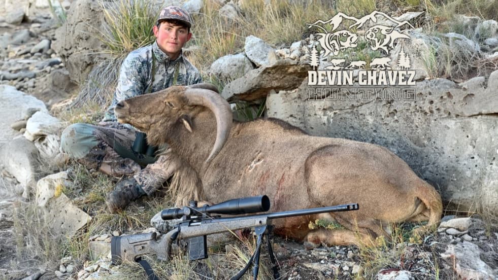 Hunter in camo next to large, dead ram with curved horns. Rifle with scope in foreground. Rocky, outdoor setting. Devin Ch...