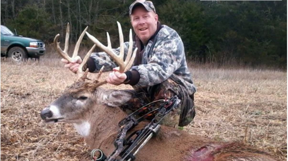 A man in camouflage kneels next to a dead deer, holding its large antlers. A bow rests nearby. A green truck is in the bac...