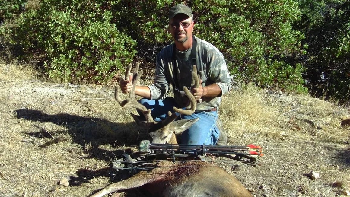 Man kneels next to a harvested deer, holding its antlers. A compound bow and arrows lie nearby. Background of dry grass a...