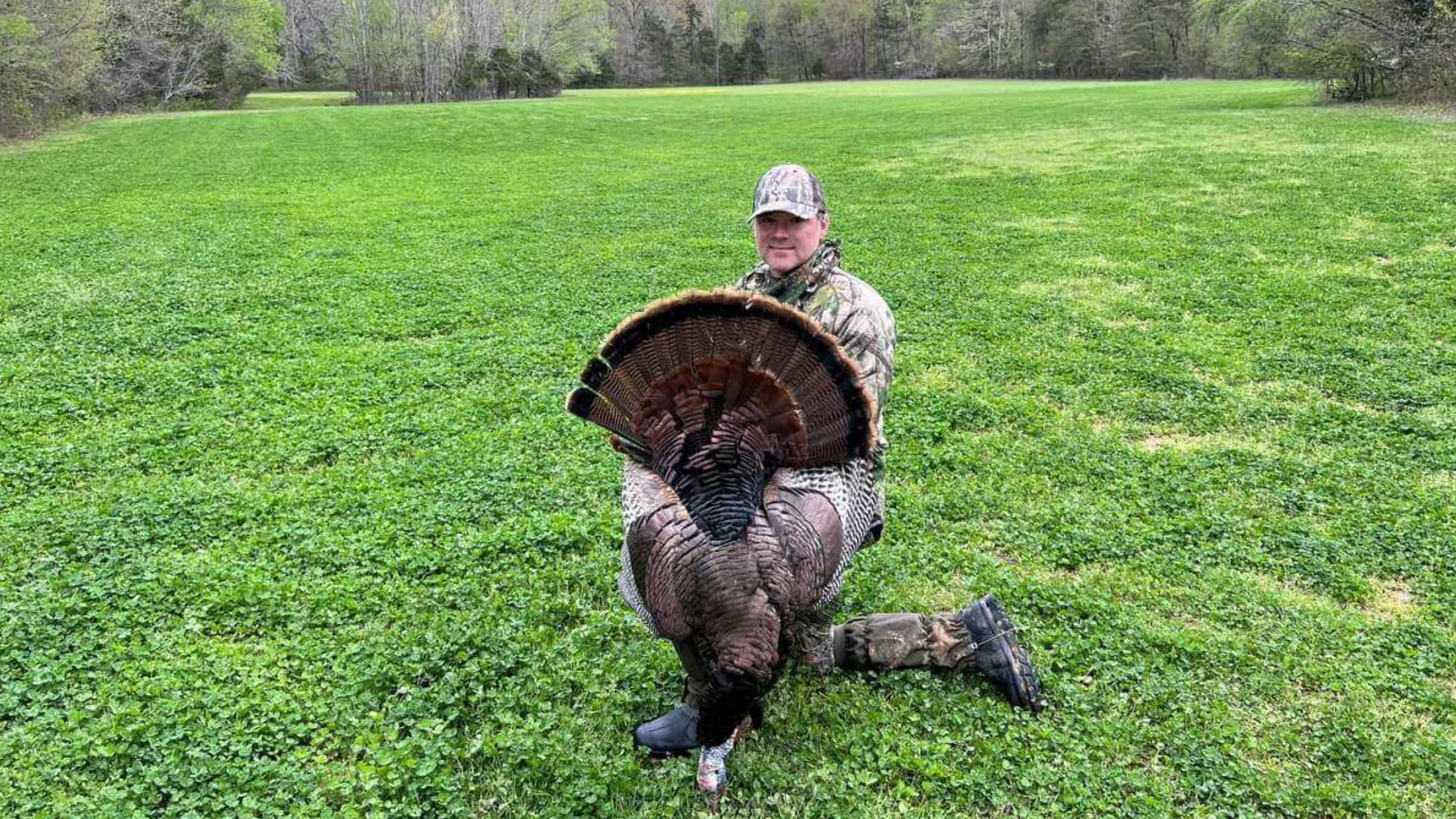 Man in camouflage kneels, holding a large wild turkey with its tail feathers fanned out. Green field and treeline in the b...