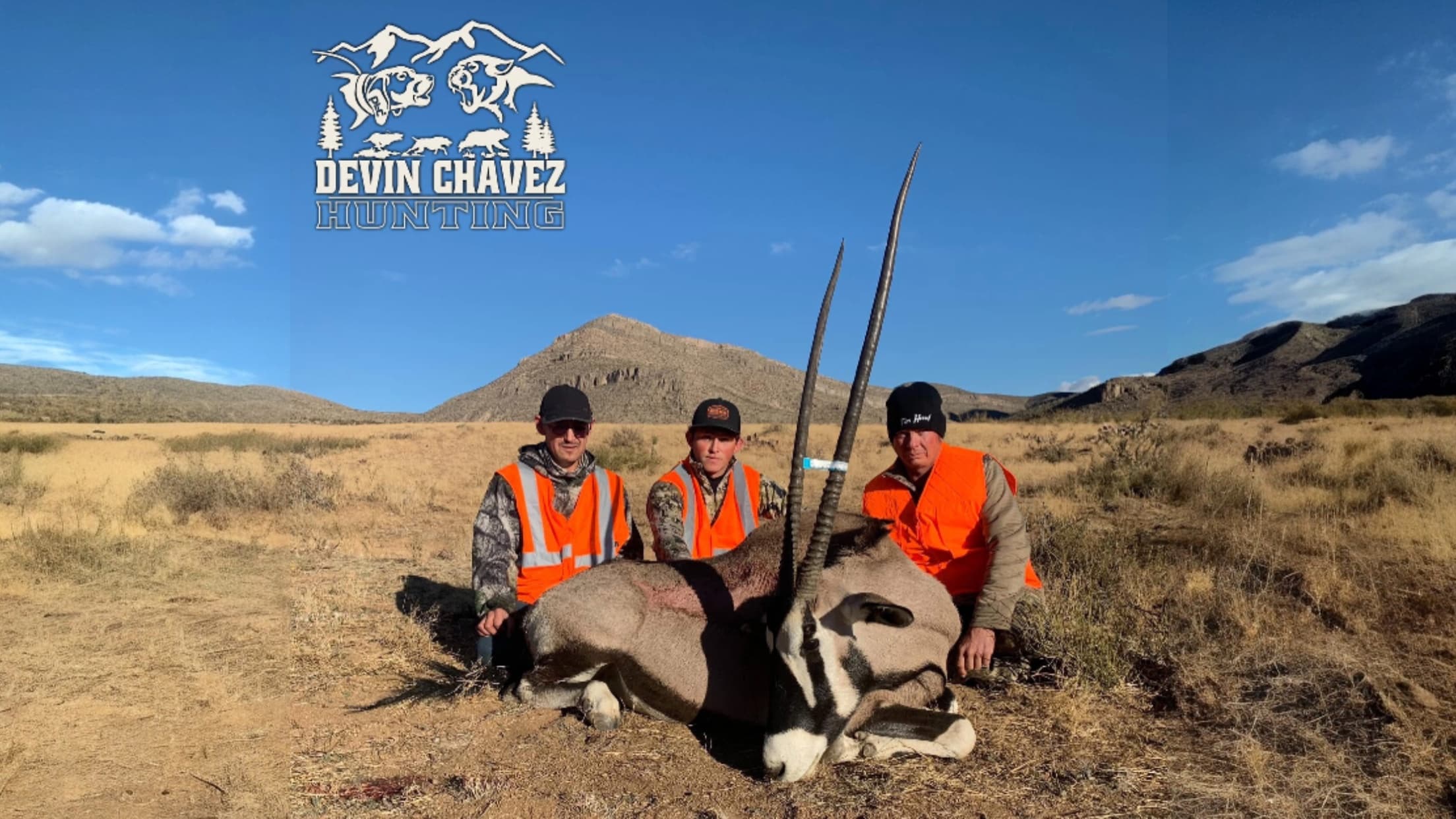 Three hunters kneel beside a deceased Oryx with long horns. All wear orange vests. Logo for Devin Chavez Hunting is at to...
