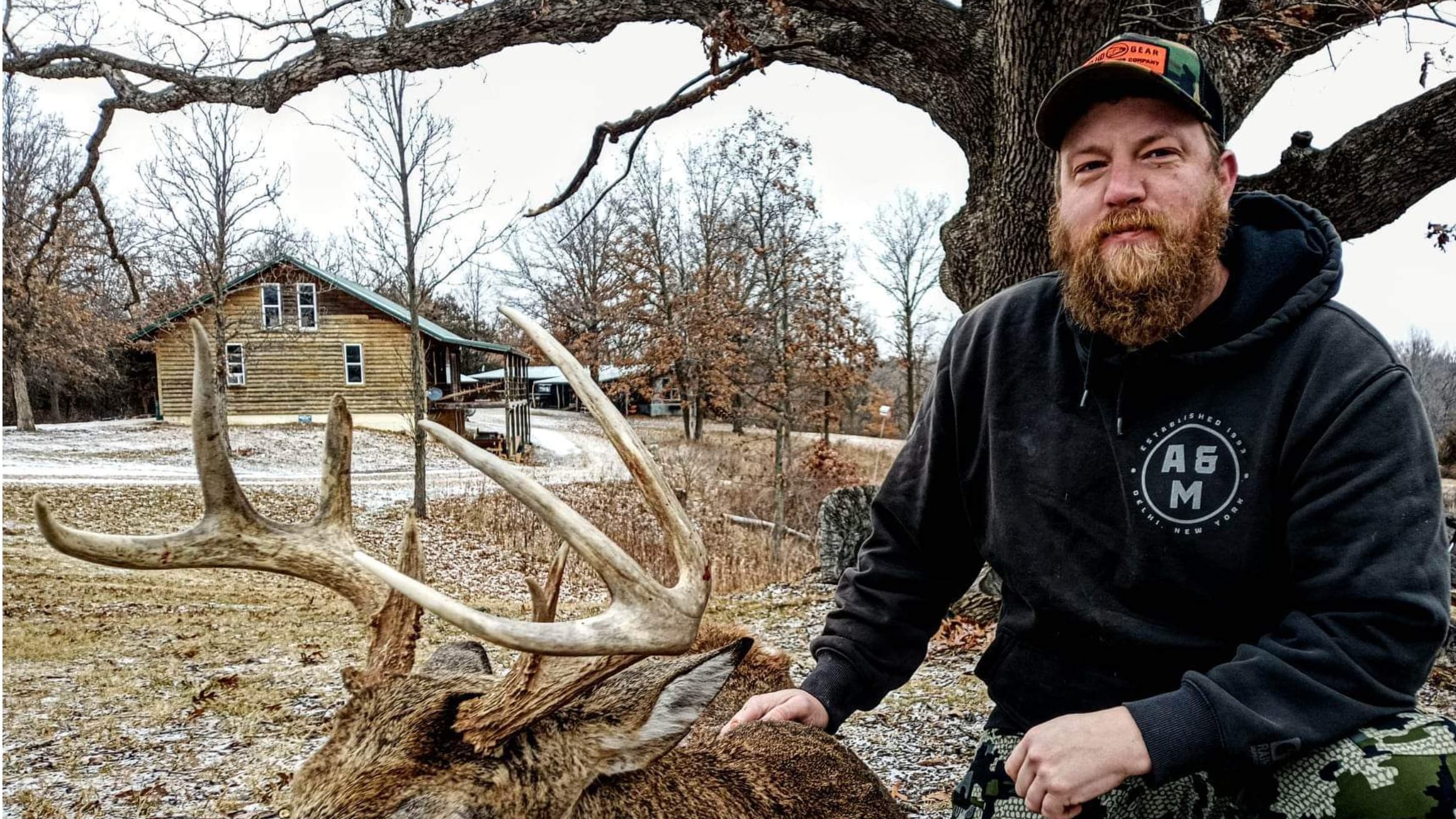 Man with large antlers, wearing a black hoodie and camouflage pants, with a log cabin in background.