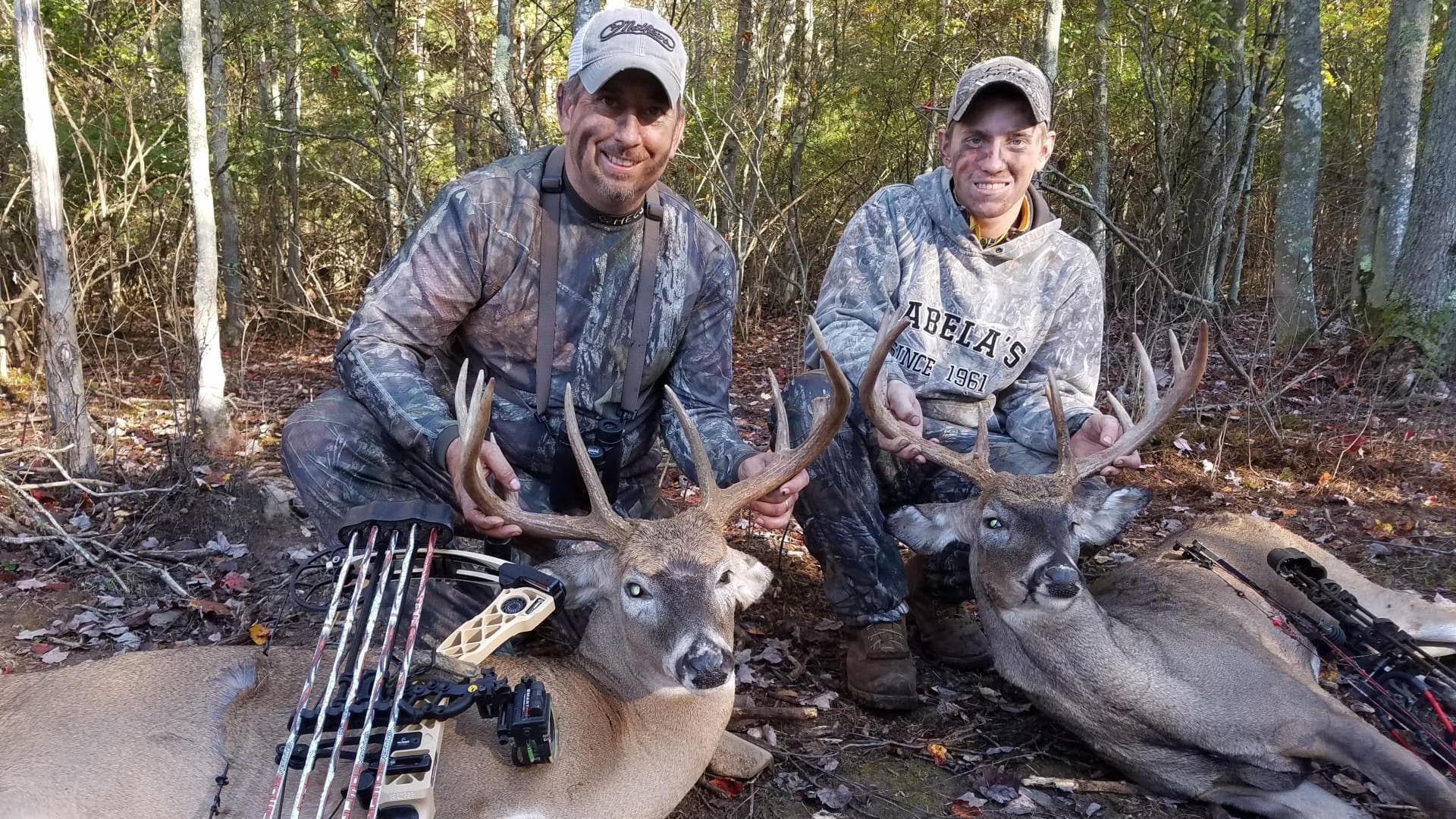 Two men in camouflage kneel, smiling, with two large deer, holding their antlers. Bows lie beside the deer in wooded area.