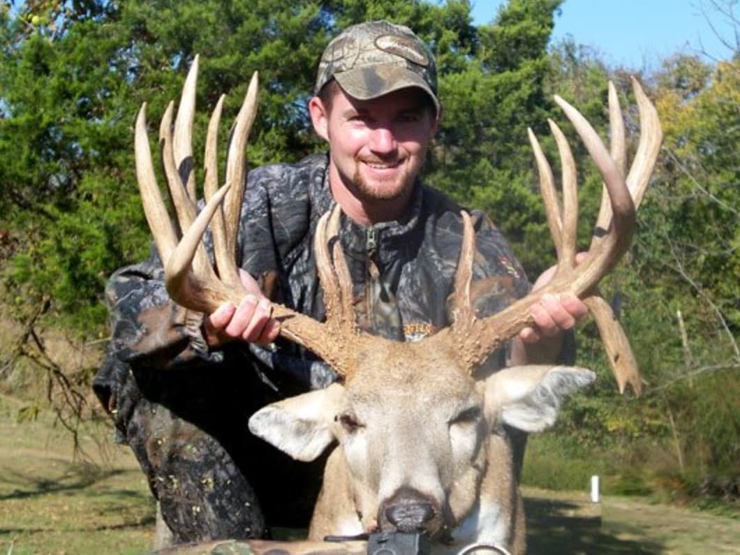 Man in camo holding massive buck with large antlers, smiling at the camera.  Deer head is visible, eyes closed. Forest bac...