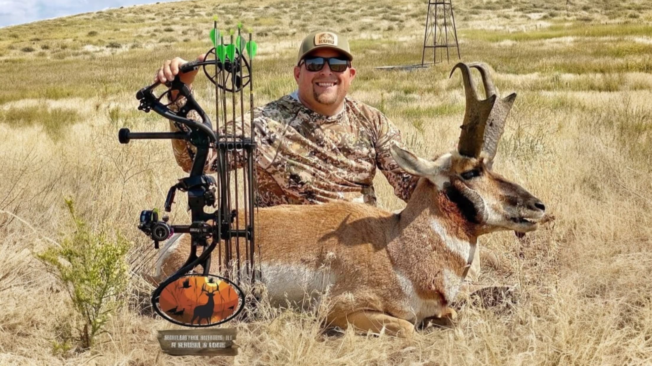 Hunter with bow poses with a pronghorn. The animal is lying in dry grass. Hunter wears camo, sunglasses, and a cap. A wind...