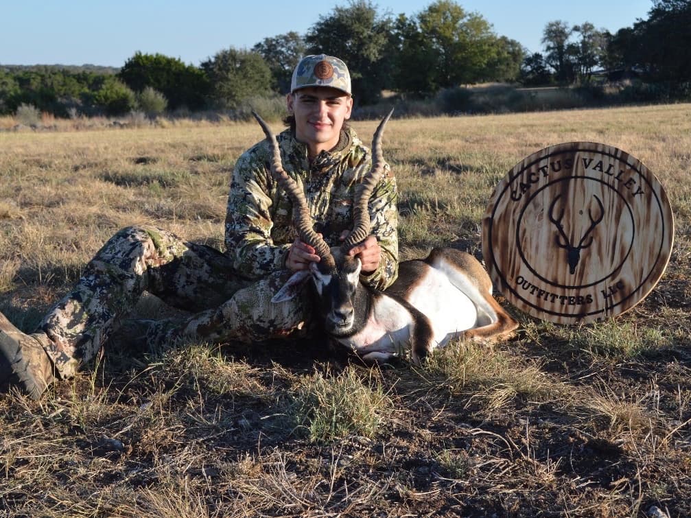 A young man in camo attire sits with a blackbuck antelope. He holds the horns, near a "Cactus Valley Outfitters" sign.