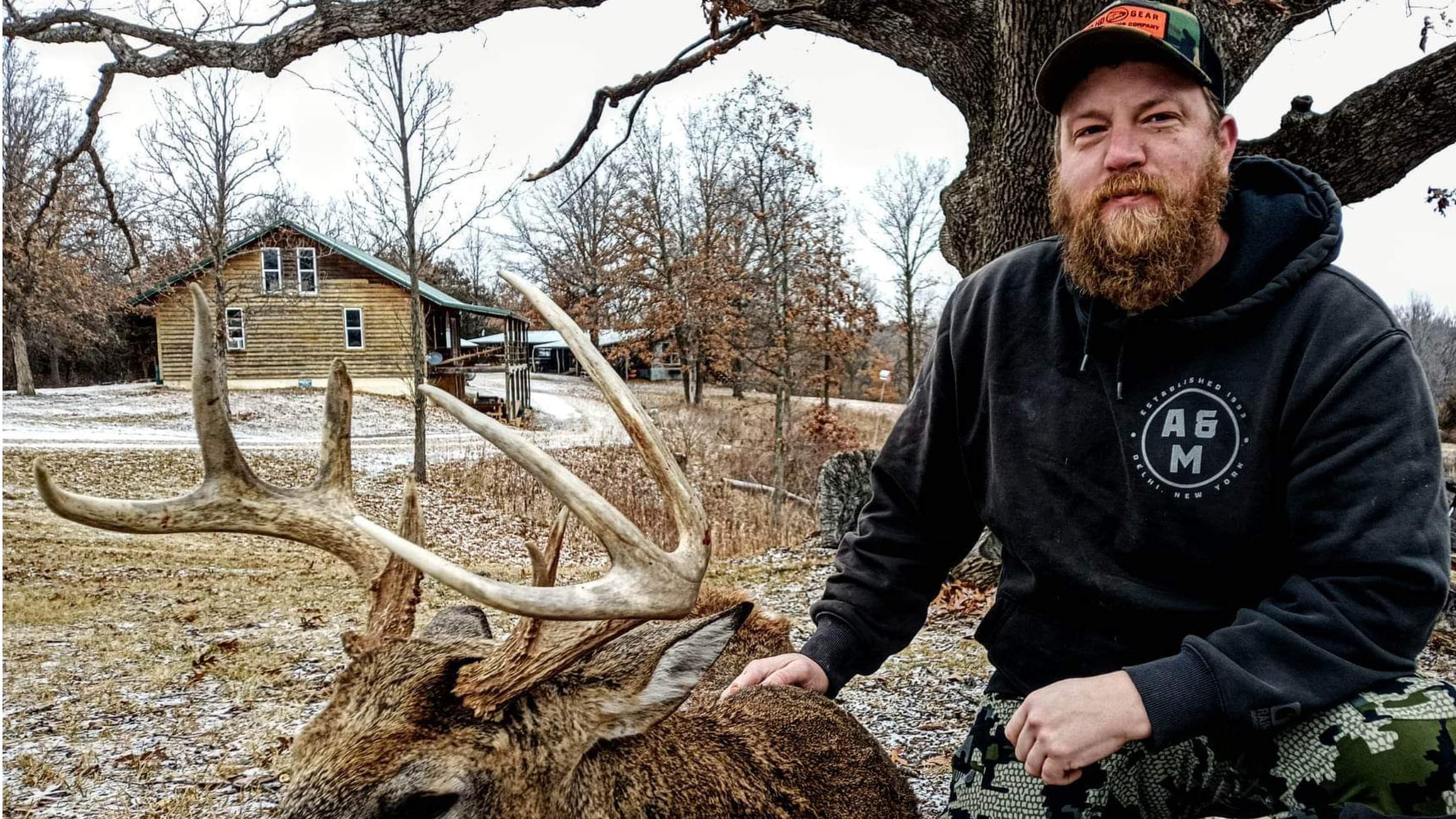 Man with beard poses next to a deer with large antlers.  A house and trees are in the background, with a grey, cloudy sky ...