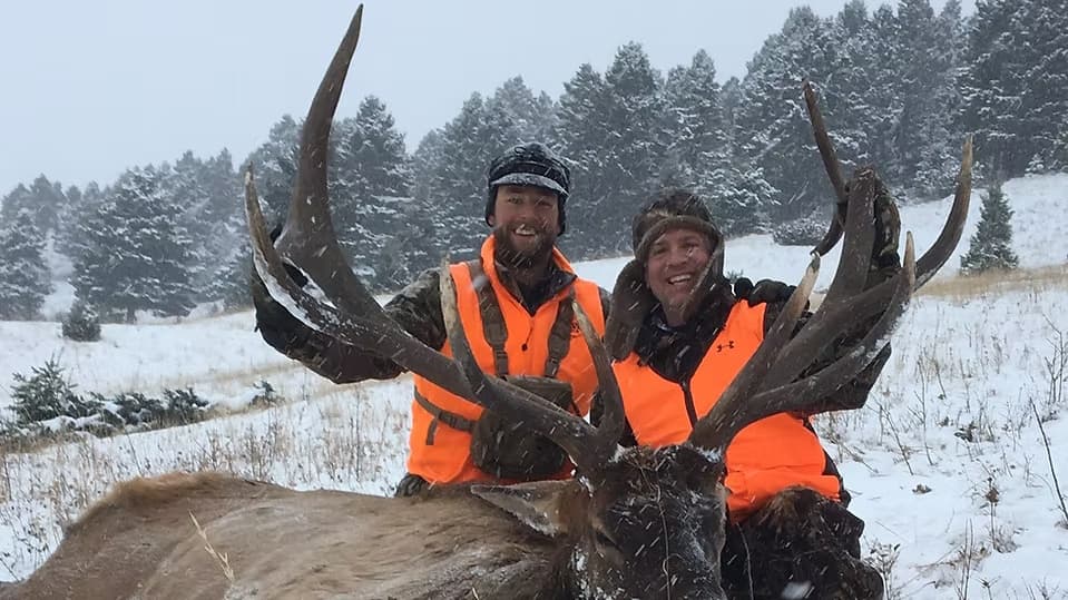 Two hunters in orange vests pose with a large elk rack in the snow, with pine trees in the background.