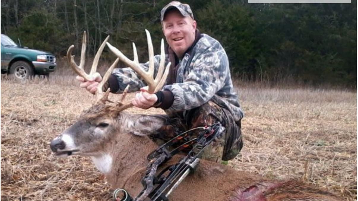 Hunter kneeling with large buck antlers, bowhunting equipment visible. Green truck in background.