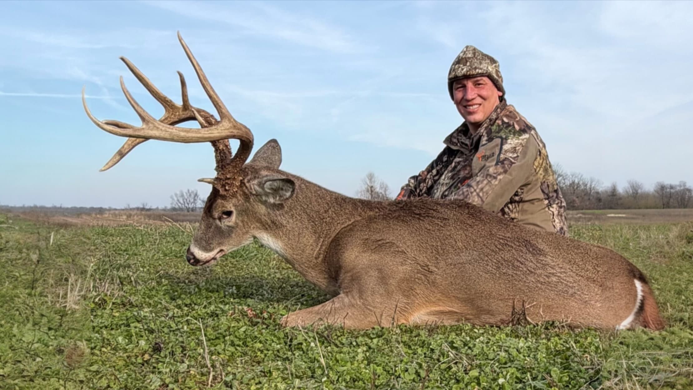Hunter with a large buck. The buck lies on green grass, with large antlers. Man smiles at the camera, wearing camo clothin...