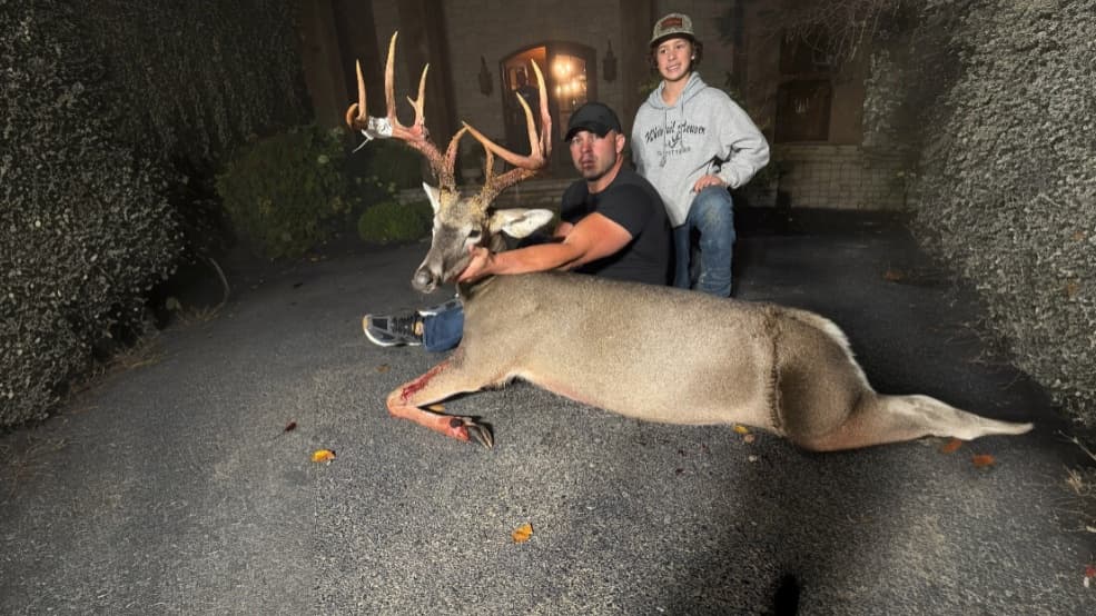 Two men and a large buck with impressive antlers on a driveway. One man kneels holding the deer's head, the other kneels b...