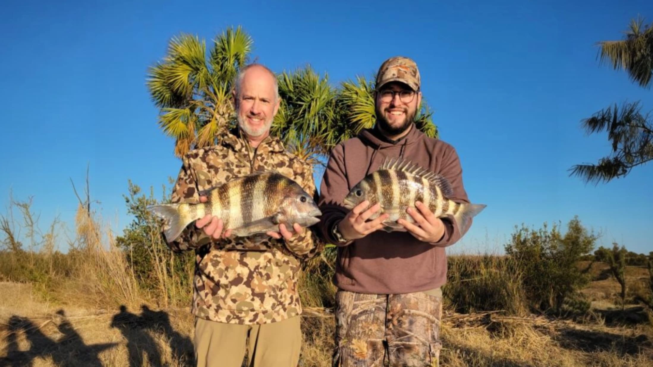 Two men holding striped fish, smiling. One in camo, one in a brown hoodie. Blue sky and palm trees in the background.