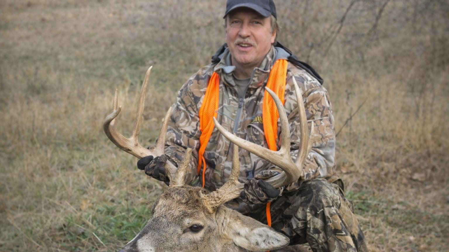 Hunter kneels with harvested buck, holding its large antlers. Man wears camo, orange vest, and a cap. Dry grass in backgro...