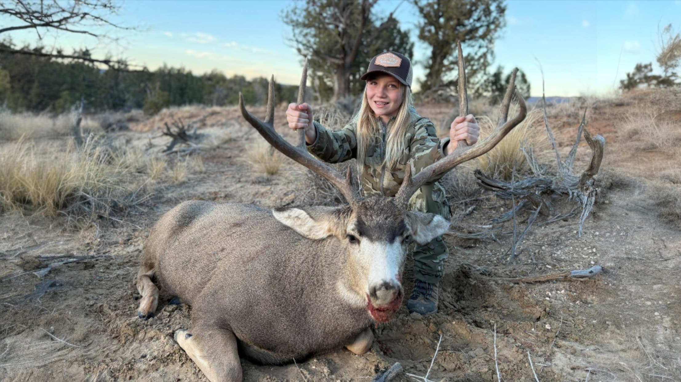 A young girl in camouflage kneels beside a large, deceased deer with large antlers. The deer lies on the ground, blood aro...