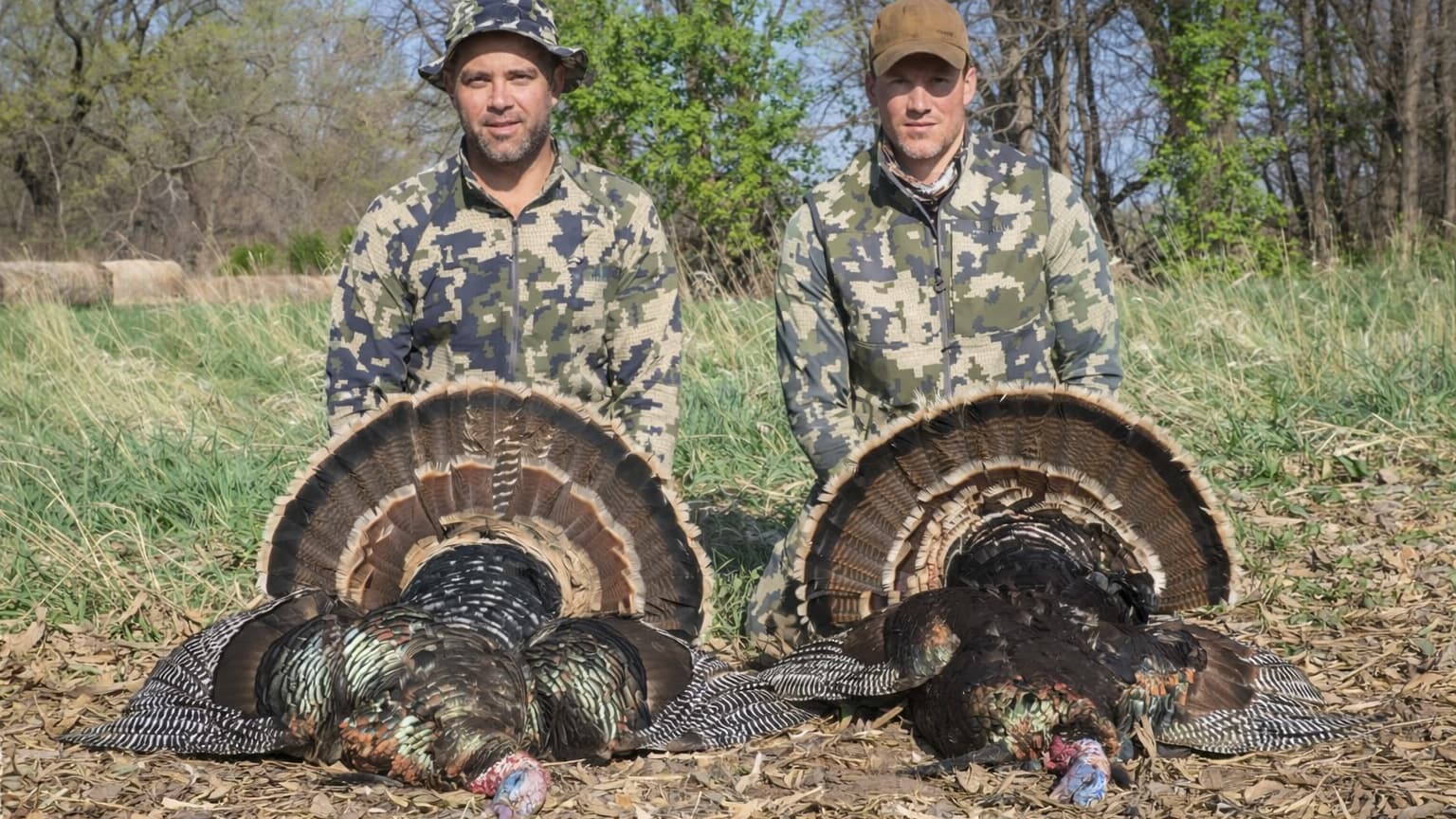 Two men in camouflage kneel behind two wild turkeys with fanned-out tail feathers. The men appear proud in a grassy field.