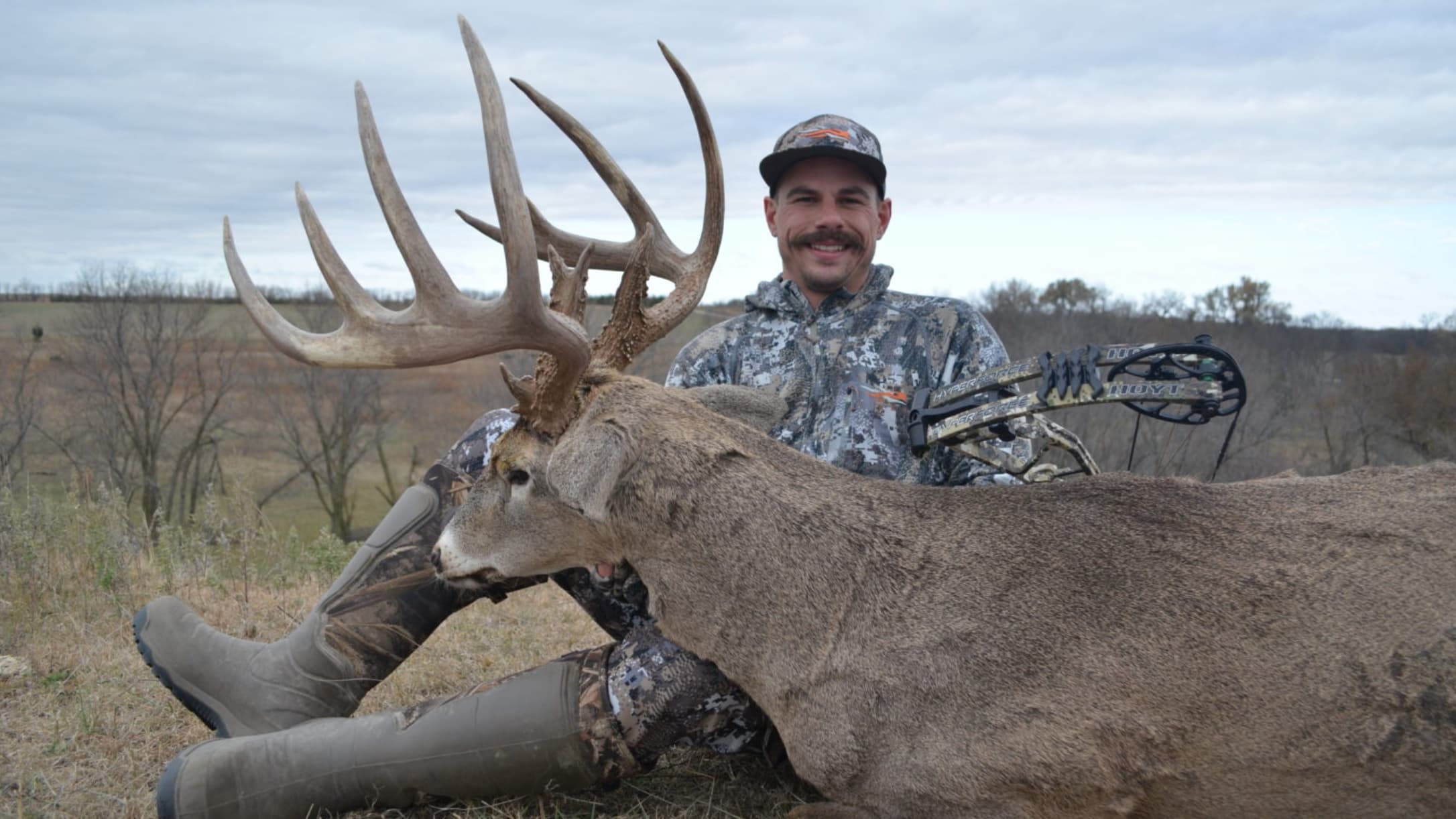 Hunter in camouflage clothing with a large buck, antler prominent. A bow sits to the side. Field and cloudy sky background.
