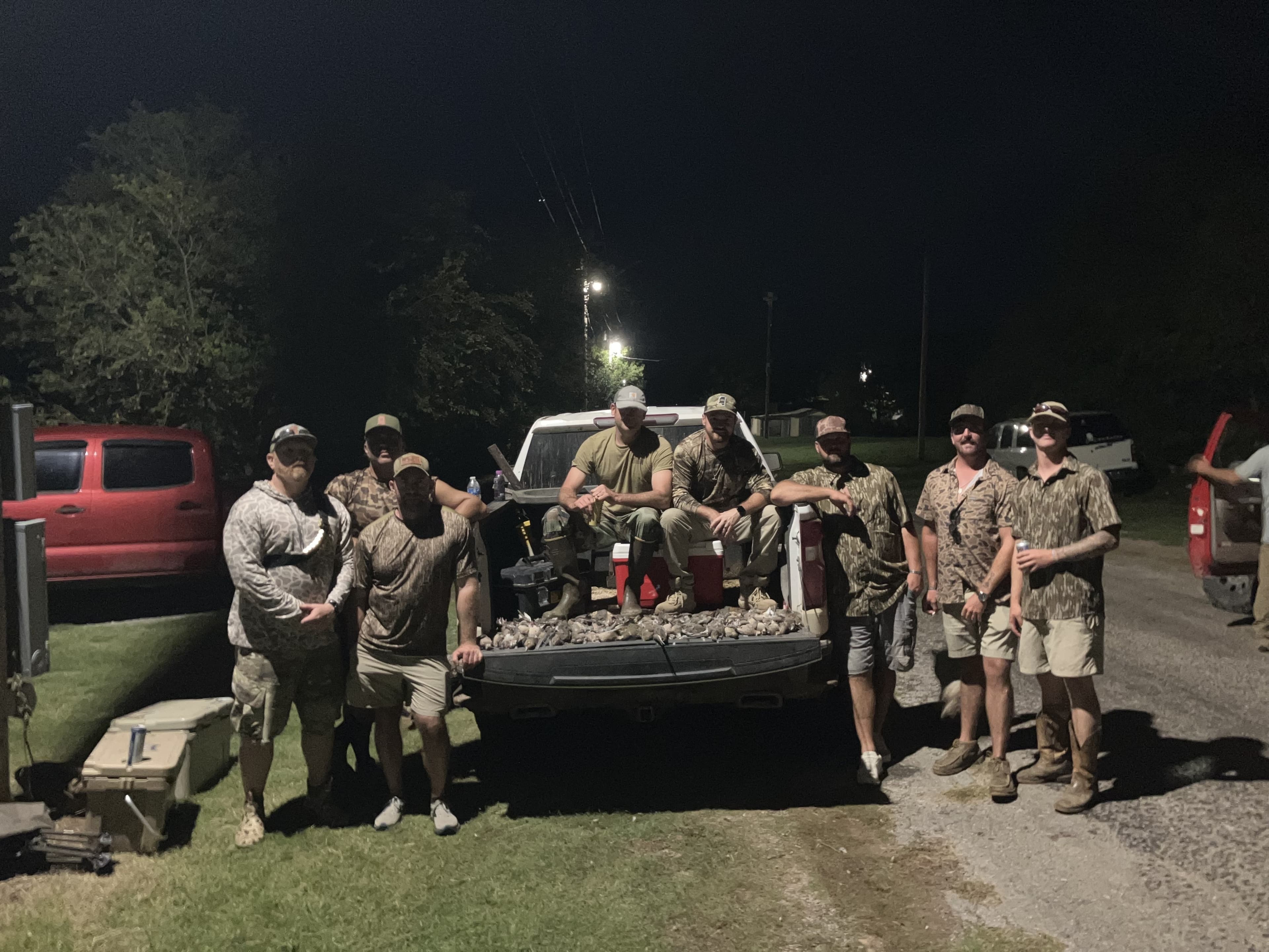 A group of men posing by a truck bed filled with hunted birds. Several wear camo and hats, under the night sky.