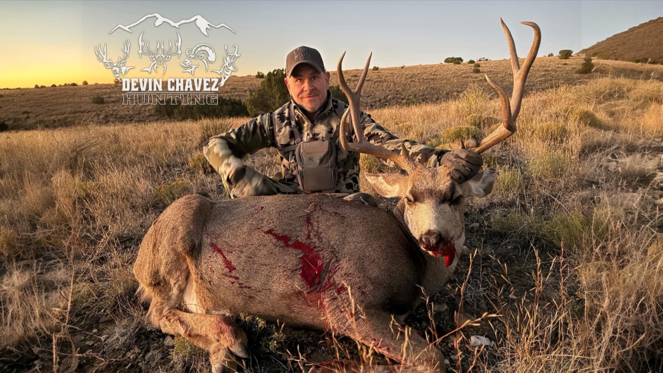 Man in camo kneels next to a large deer with large antlers. Deer has visible wounds. Field and setting sun in the background.