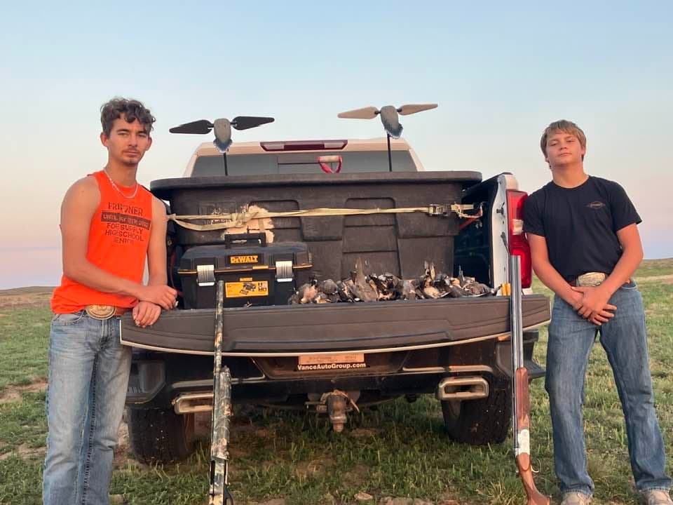 Two young men standing beside a truck bed filled with harvested birds and hunting gear. One holds a shotgun. Evening sky.