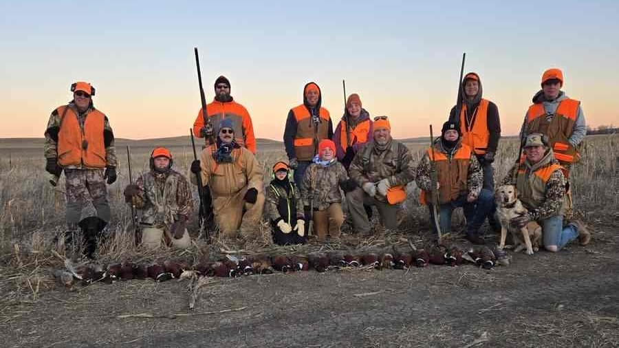 A large group of hunters, including children, pose with shotguns and a dog, behind a row of hunted pheasants.