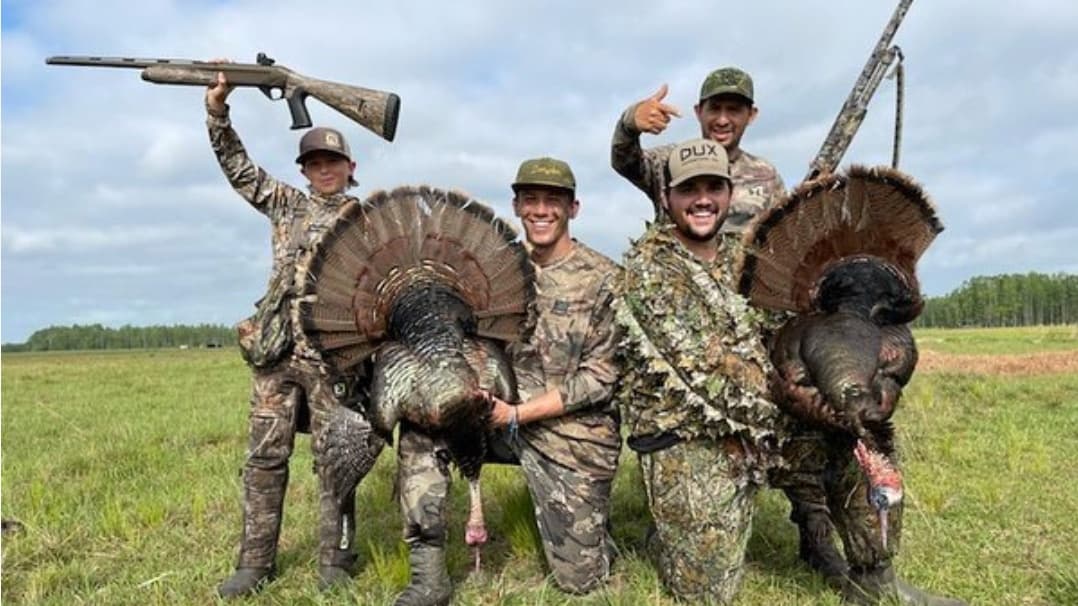 Five men in camouflage with shotguns pose with harvested turkeys. One holds a gun up, others hold the birds, smiling, in a...
