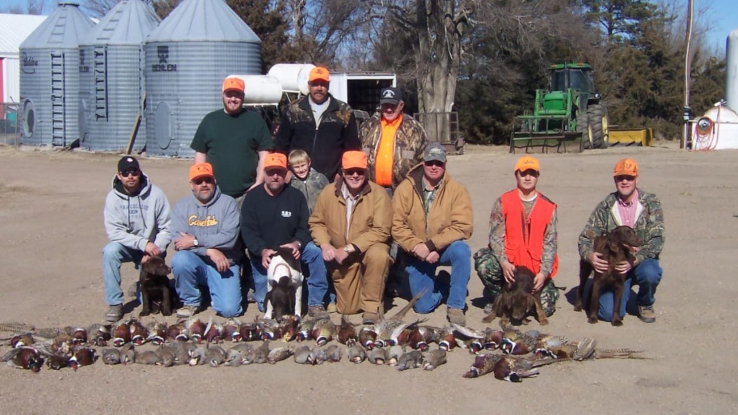 Group of men with dogs posing in front of a harvest of birds. Men wear orange hats, farm silos in background.