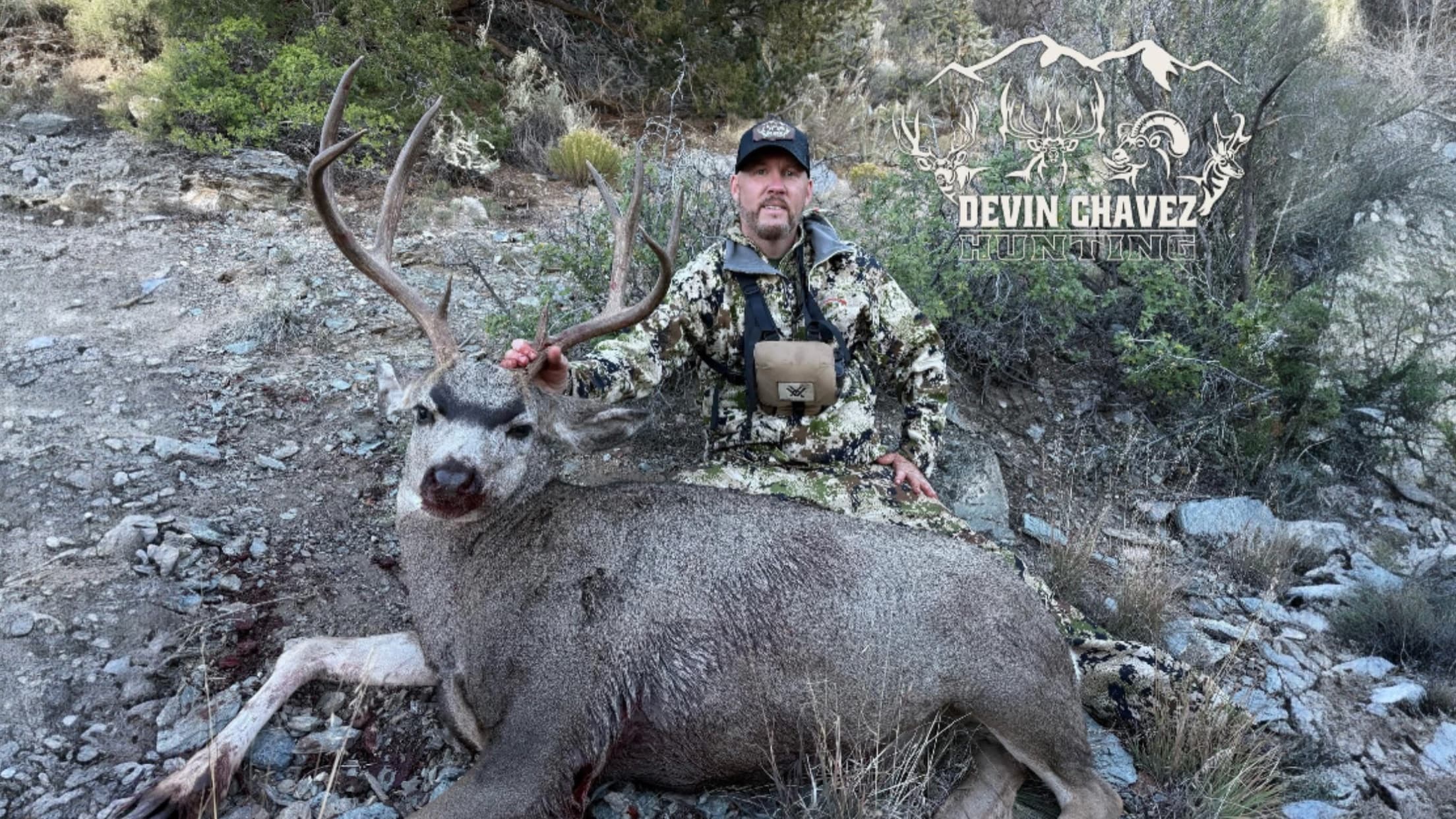 Hunter in camo gear kneels next to a large buck with impressive antlers. Buck is lying on rocky ground, with blood visible.